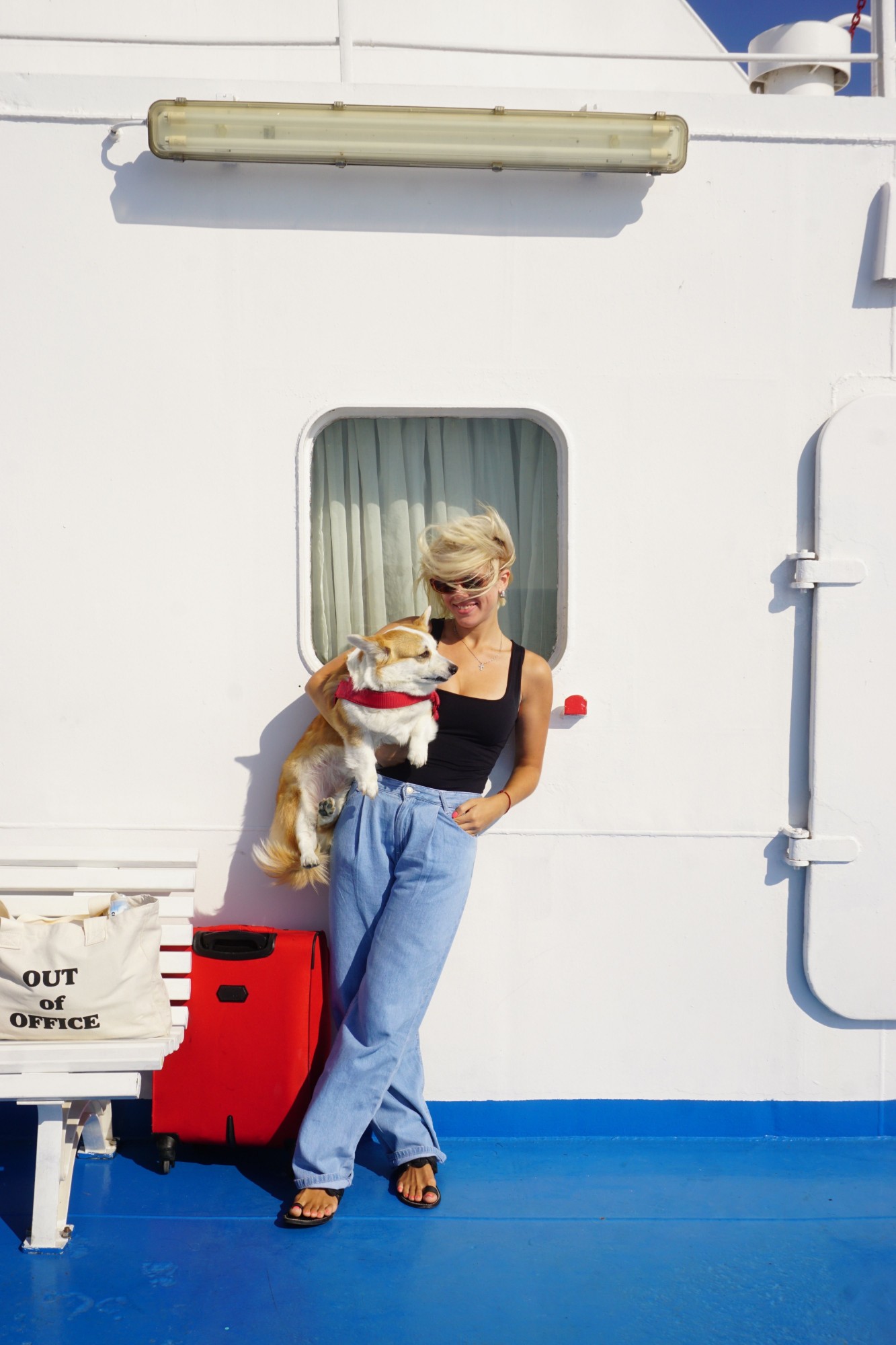 A woman holding a corgi in a red harness on a sunny ferry deck, next to a red suitcase